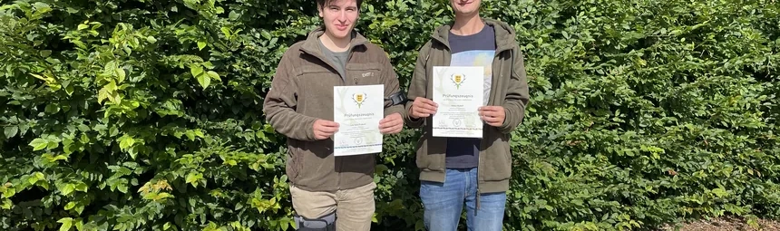 Zwei Absolventen stehen vor einer Hecke und halten ihr Prüfungszeugnis in die Kamera