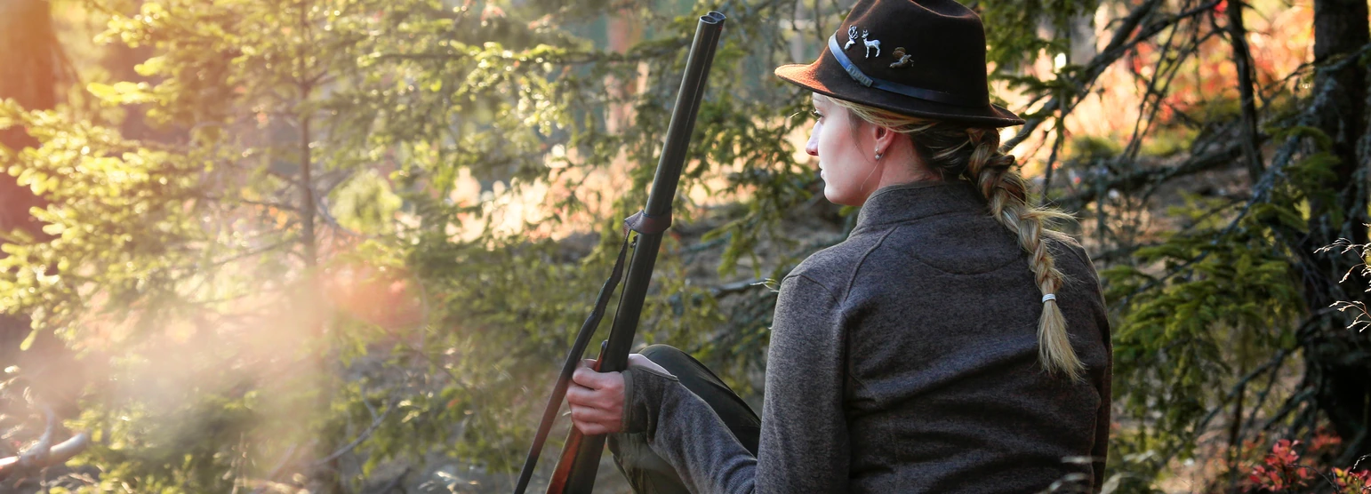 Jägerin sitzt am Hang im herbstlichen Wald und blickt ins Gegenlicht, das Gewehr aufrecht neben sich
