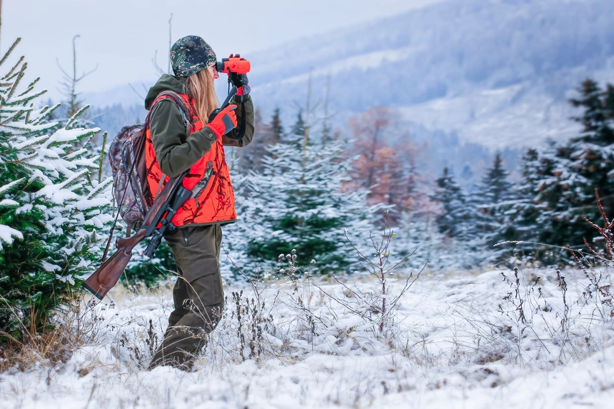Jägerin mit Fernglas und Büchse steht im verschneiten Nadelwald