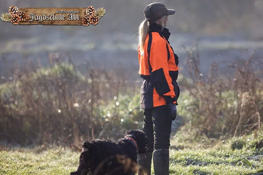 Jägerin in Signalkleidung steht mit ihrem Hund am Rand einer Wiese