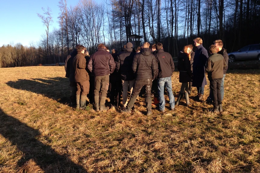 Eine Gruppe in Jagdkleidung steht im Halbkreis auf einer Wiese am Waldrand, im Hintergrund eine Kanzel zwischen kahlen Bäumen